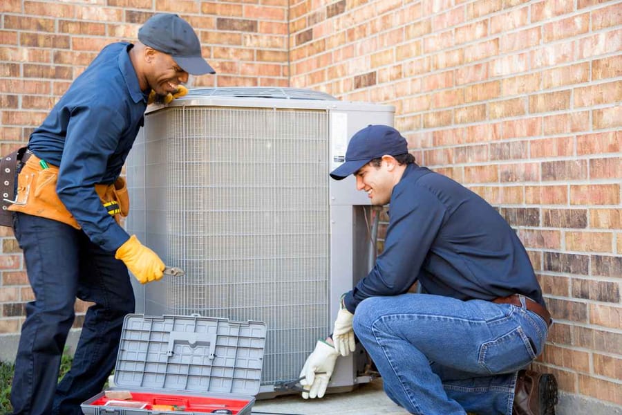 Two HVAC technicians working on an outdoor system. They are both wearing blue work shirts and smiling as they do the job.