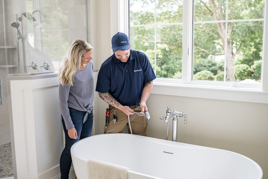 Plumber in a blue uniform fixes a bathtub faucet while a woman watches in a bright bathroom with large windows.