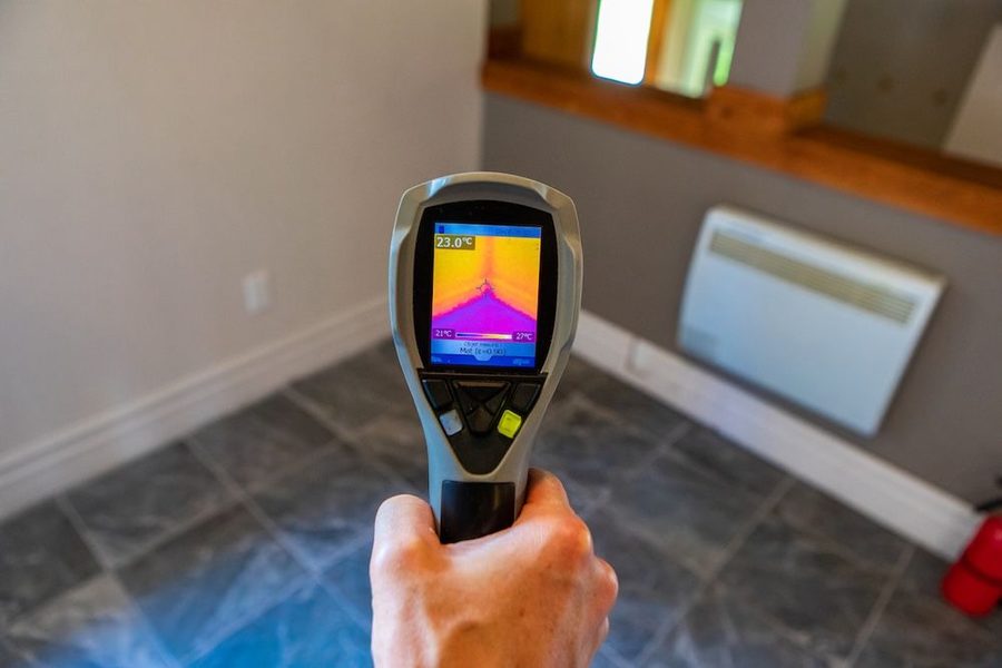 Handheld thermal imaging camera displaying a colorful heat map of the floor in an indoor room, held in a person’s hand