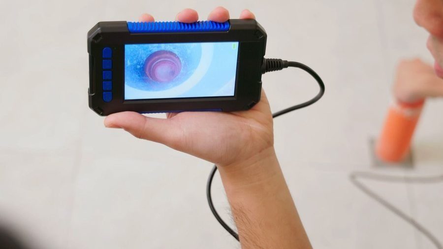 Handheld inspection camera with a blue-trim housing showing a live feed of a pipe interior on its screen, held at arm's length by a person