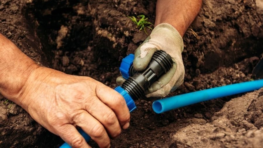 Close-up of a gloved and bare hand joining a blue PVC pipe with a connector in a dirt trench while working outdoors.